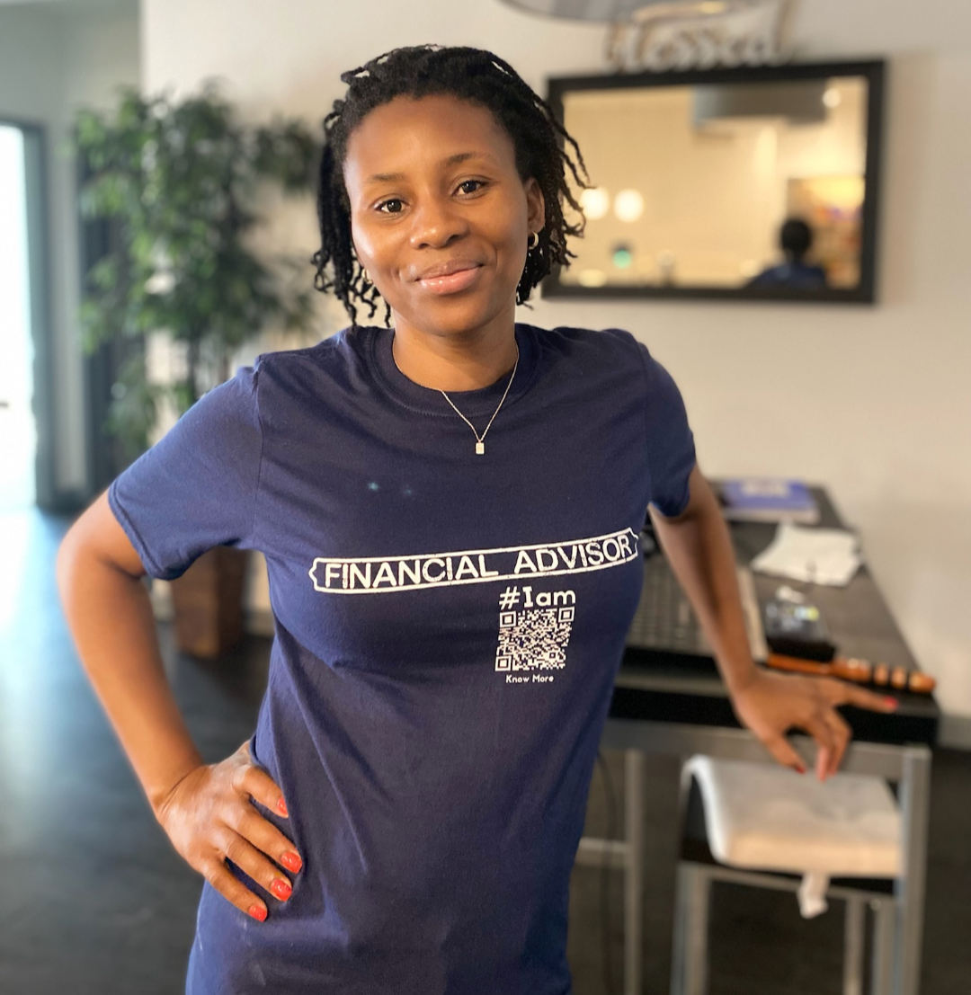 An African American woman is standing at a desk, wearing a navy blue CLAIM It Tee™. The CLAIM™ reads {FINANCIAL ADVISOR} #Iam.