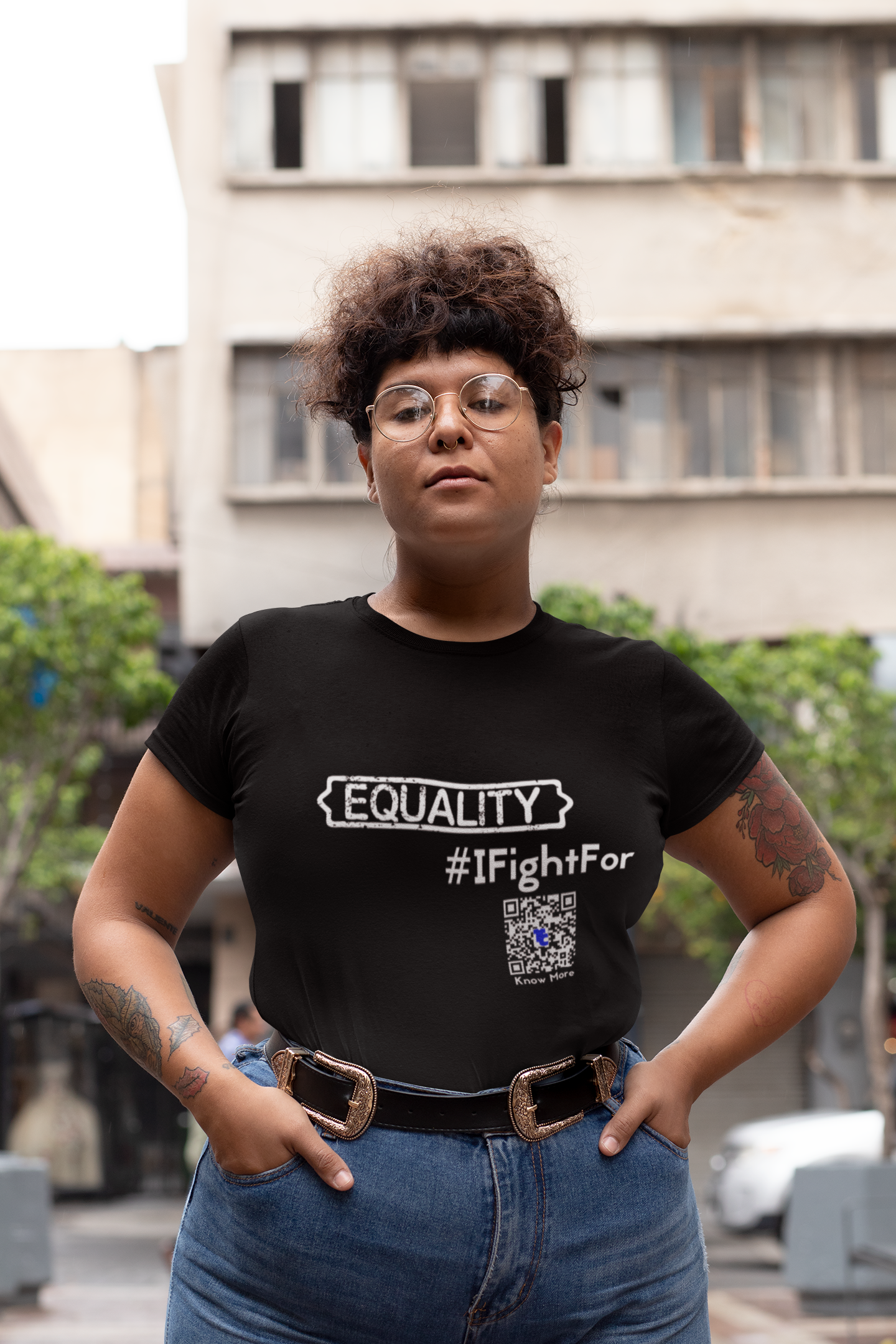 A female presenting person, in jeans and a black CLAIM It Tee™ is standing on a city street with a serious look on her face. The CLAIM™ reads {EQUALITY} #IFightFor