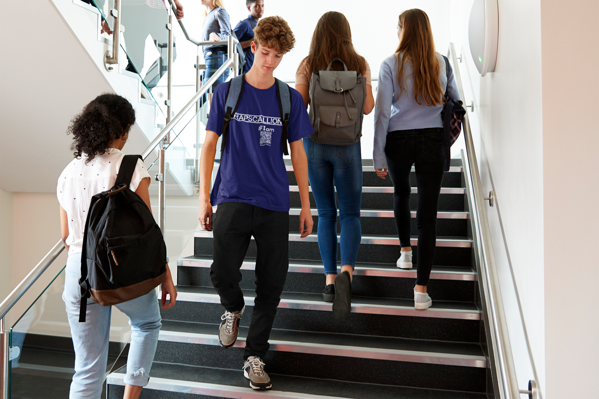Teenage students going up & down stairs in the stairwell of a modern looking school or public space. A male presenting young person, wearing black jeans & a navy blue CLAIM It Tee™  with a CLAIM that reads {RAPSCALLION} #Iam.