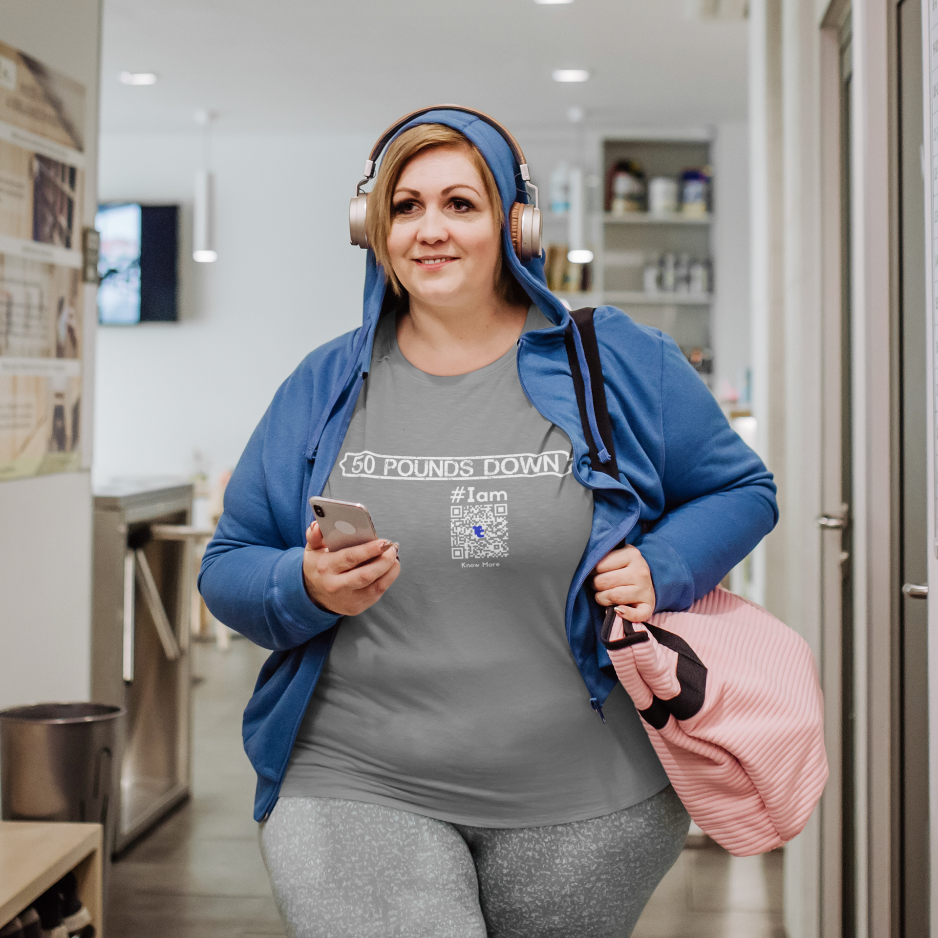 A female presenting, full figured, person is TeeCLAIMING™ as they walk indoors with a gym bag & headphones. They're wearing a gray CLAIM It Tee™ with a CLAIM™ that reads {50 POUNDS DOWN} #Iam.