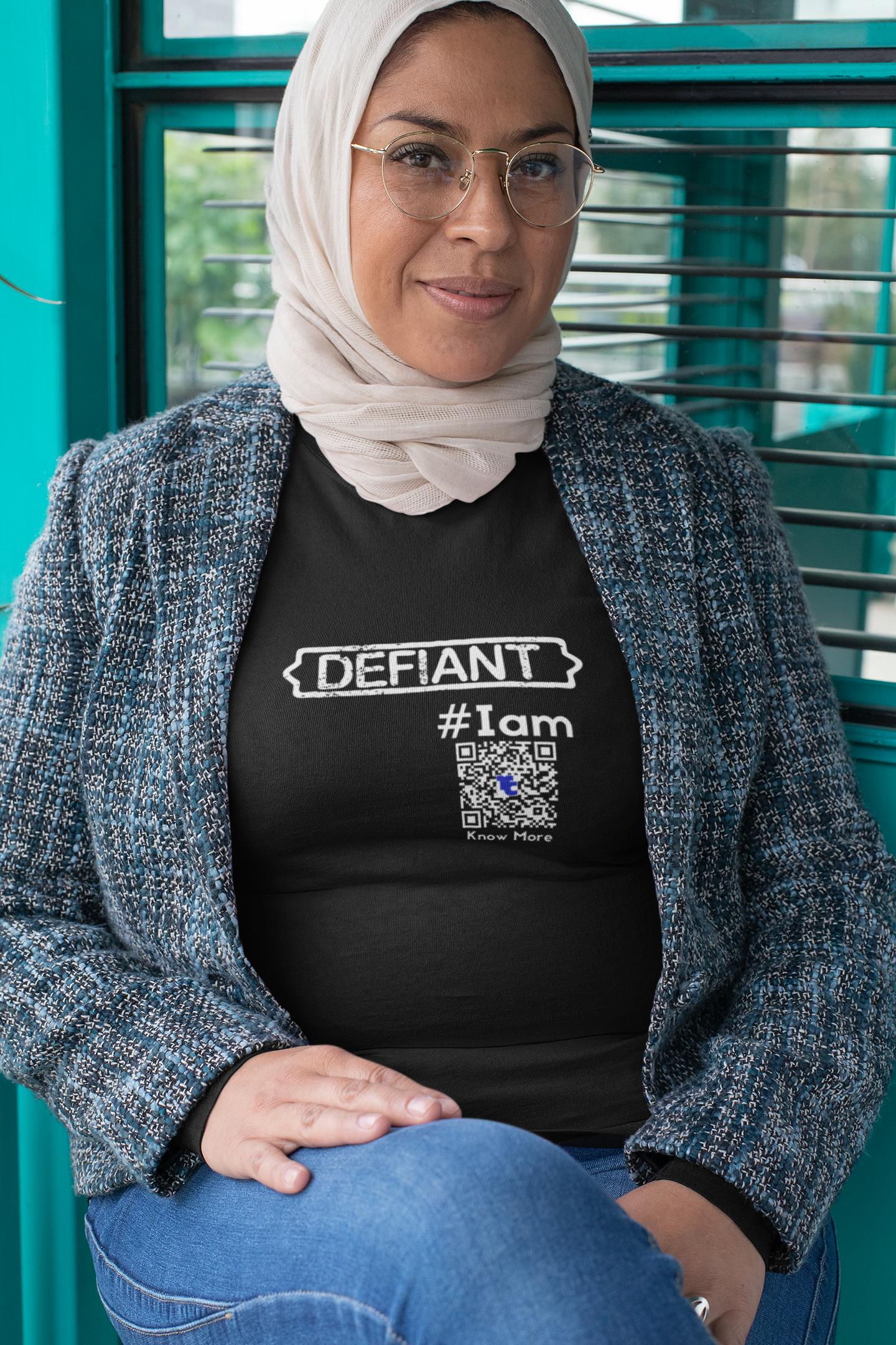 Female presenting person seated outside, during the day, in blue jeans wearing a black CLAIM It Tee™ that reads {DEFIANT} #Iam. She is wearing a blue and black tailored jacket over it and a cream head scarf covering her dark hair. 