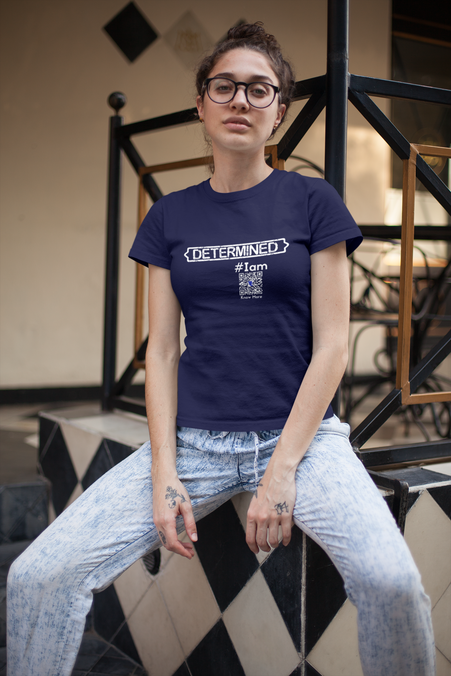 A female presenting person wearing acid washed jeans, glasses, and a navy blue, short sleeve, cotton, crew neck tee with the TeeCLAIM Determined #Iam and unique QRcode for that shirt's digital share space.