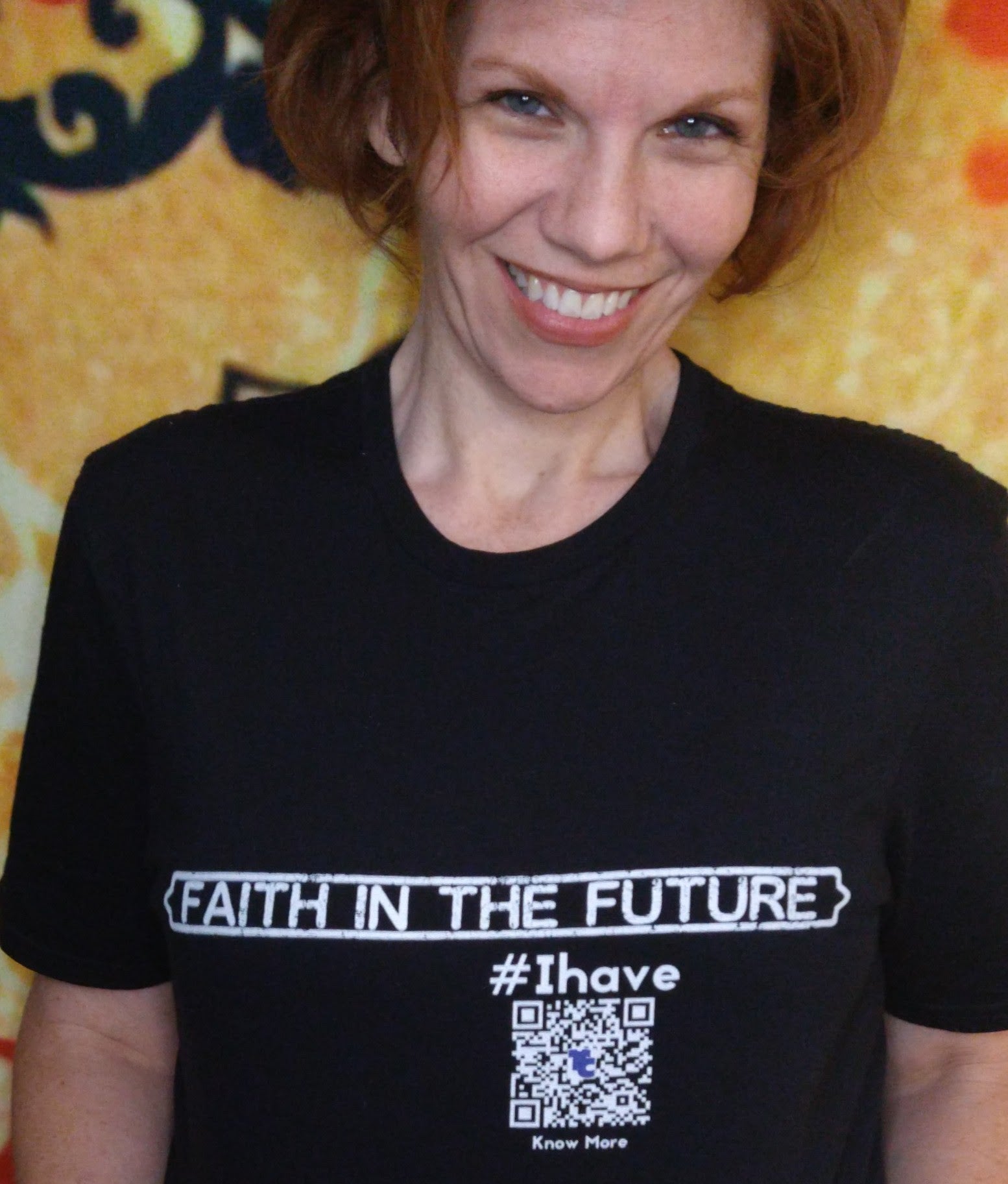 A close up picture of a smiling, female presenting, woman. She is wearing a black CLAIM It Tee™. The CLAIM™ reads {FAITH IN THE FUTURE} #Ihave. The Share Space™ can be accessed by reading the QRcode on the shirt.