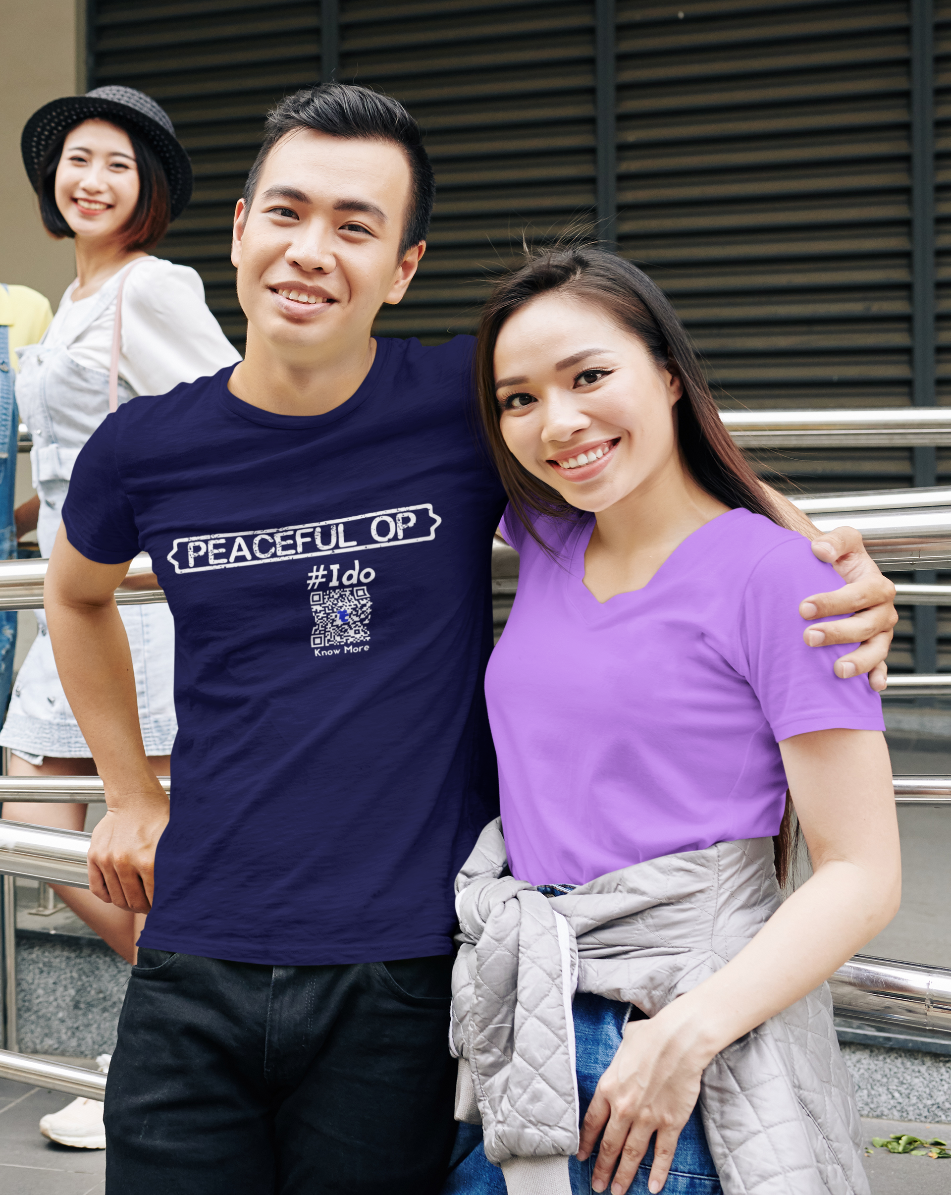 A heterosexual Asian couple are standing next to each other with one arm around the other one. They're smiling. They're outside and looking at the camera. She is wearing a solid t-shirt. He is wearing a CLAIM It Tee that says Peaceful Op #Ido