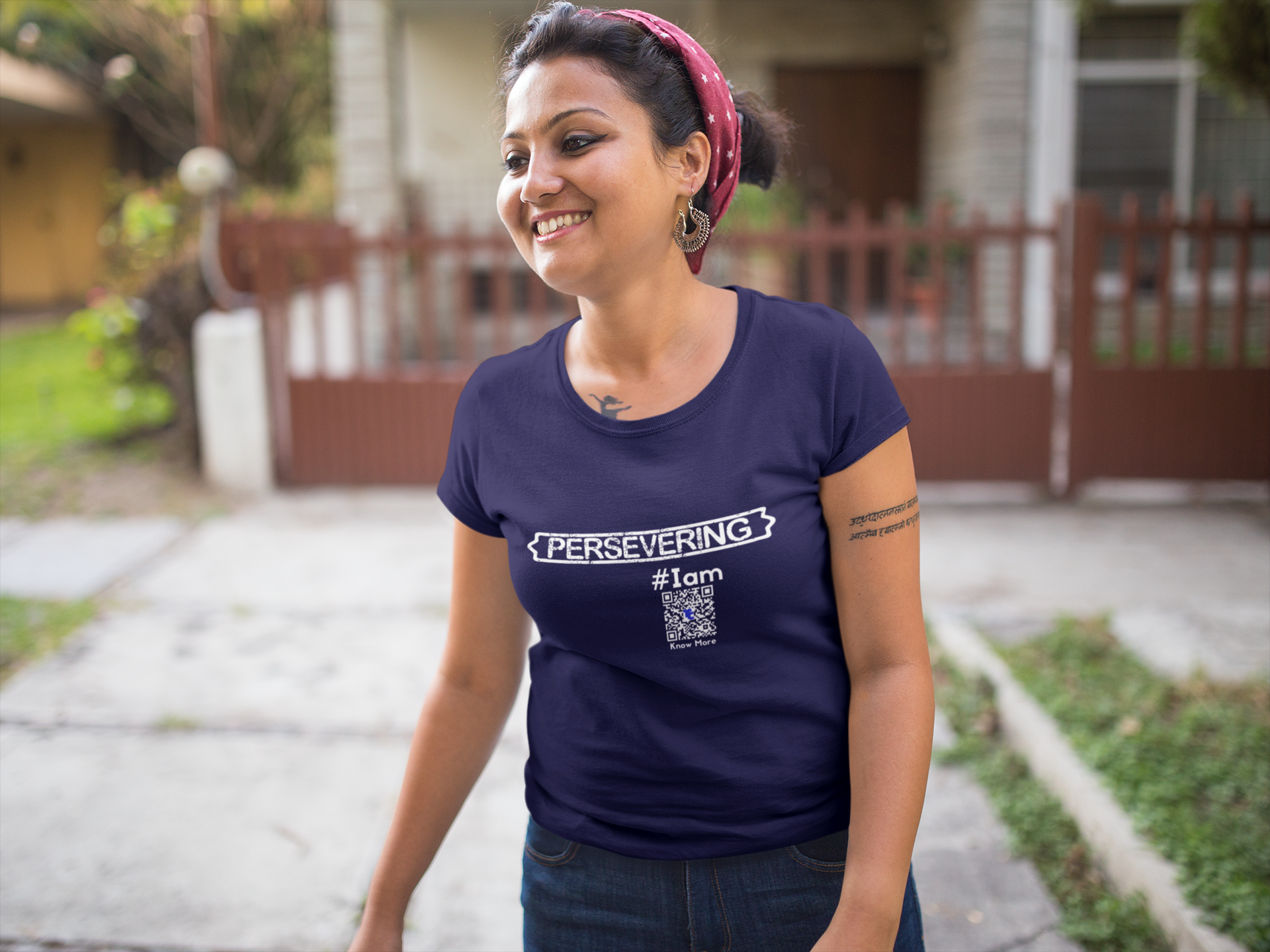 A Female presenting person is standing outside of a suburban home in a navy blue CLAIM It Tee. The TeeCLAIM is {PERSEVERING} #Iam. She's wearing blue jeans with her hair pulled up in a bun.