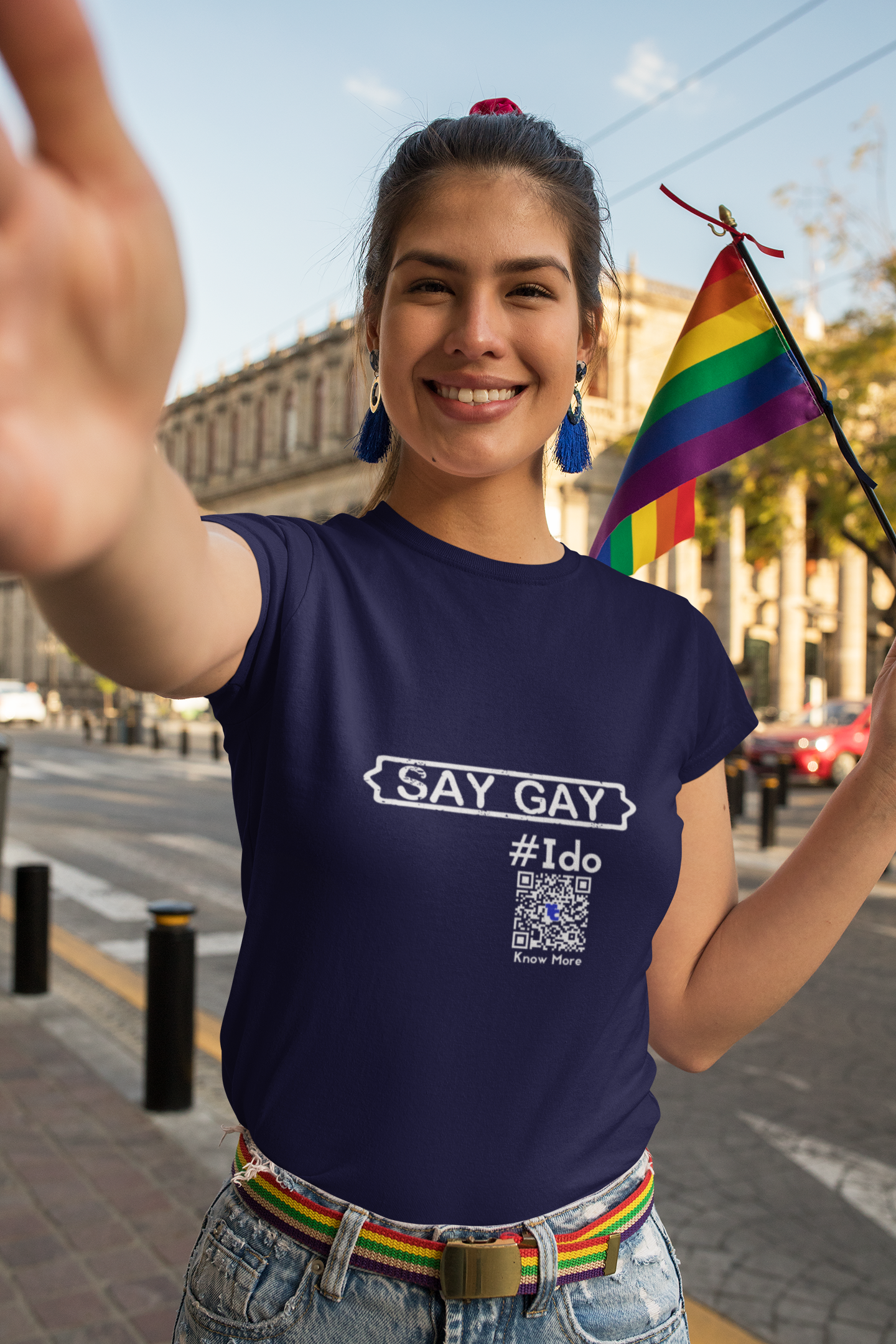 A person is walking on a city street, smiling, holding a rainbow pride flag looking directly at the camera. They're wearing a navy blue CLAIM It Tee™.  The CLAIM™ reads {SAY GAY} #Ido. The qr code is easy to read.
