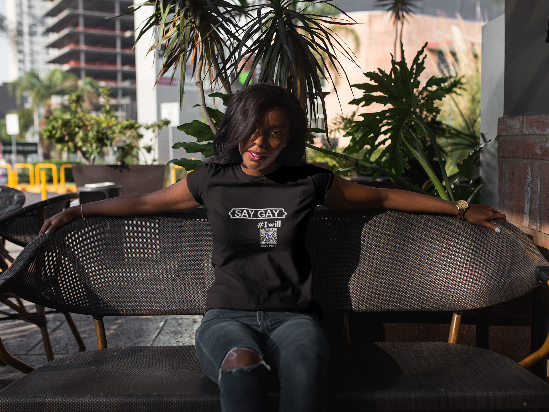 A person sitting on a chair with open arms by themselves looking at the camera. They're wearing a black short sleeve CLAIM It Tee™.  The CLAIM™ reads {SAY GAY} #Iwill. The qr code is easy to read.