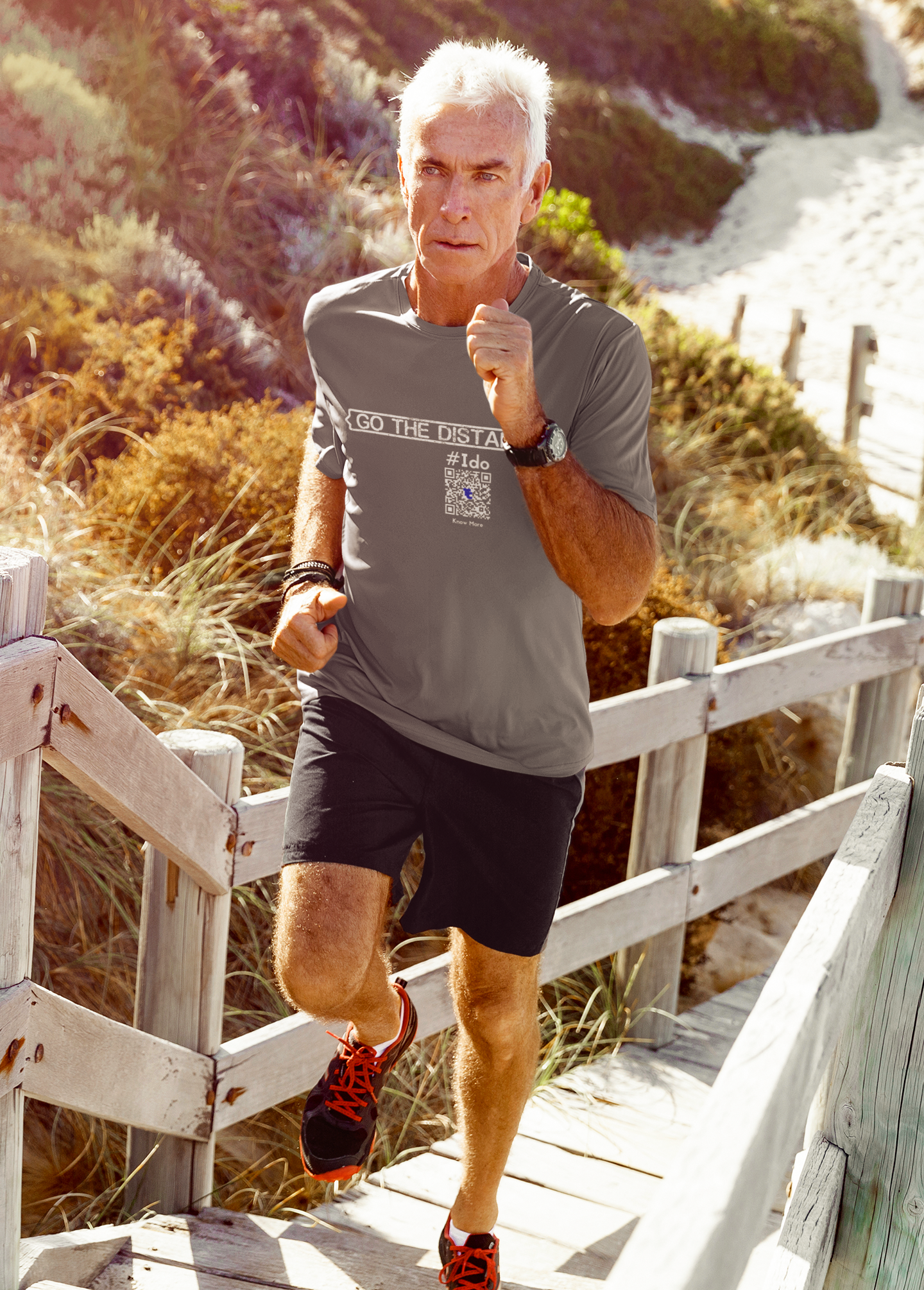 A 60+ year old male is running up a dock over sand dunes. He is wearing Black shorts, sneakers, & a gray CLAIM It Tee™. The CLAIM™ reads, {GO THE DISTANCE} #Ido.