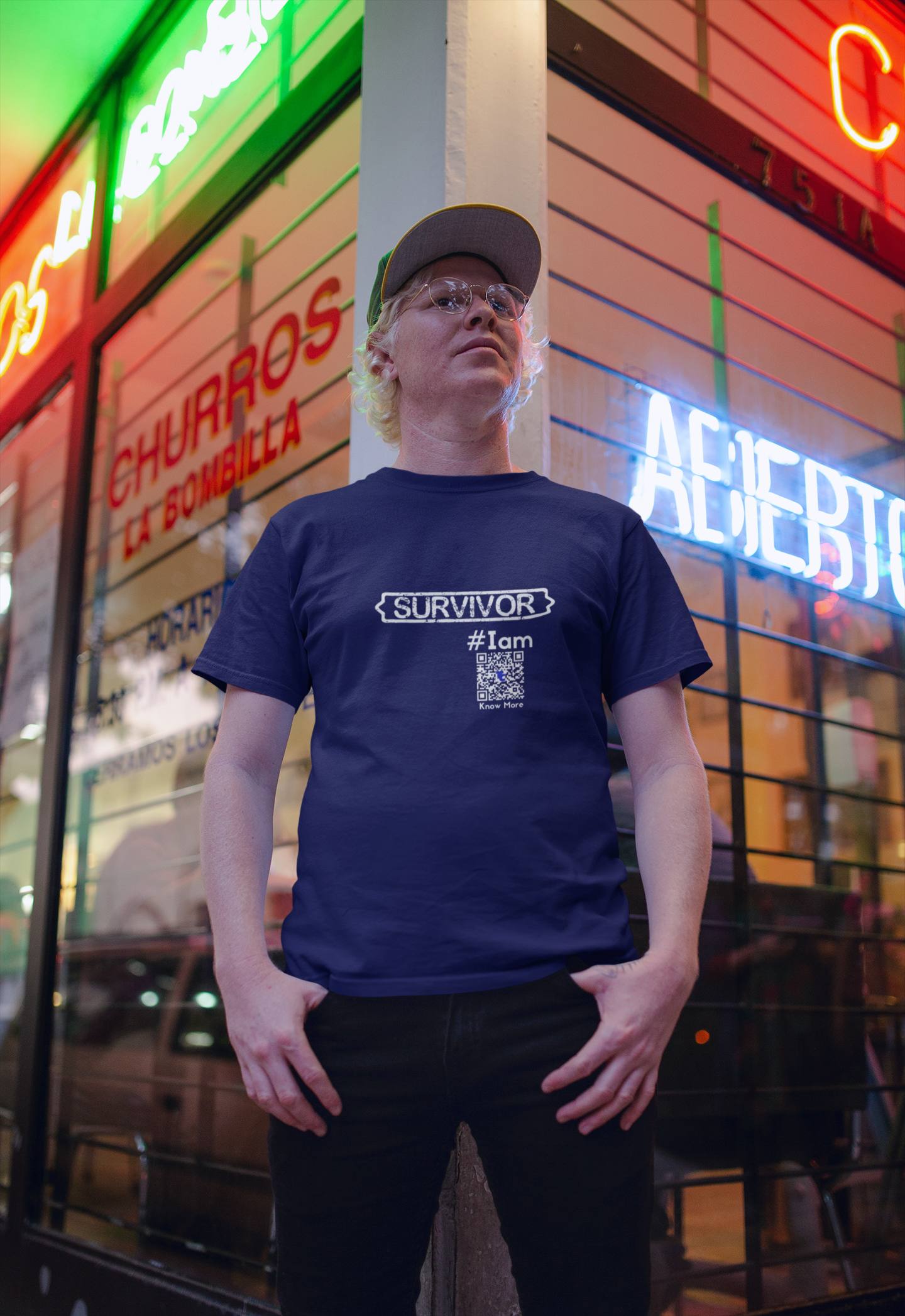 A male presenting person is standing in front a business with neon signage. They are wearing a navy blue CLAIM It Tee™. The CLAIM reads {SURVIVOR} #Iam. 
