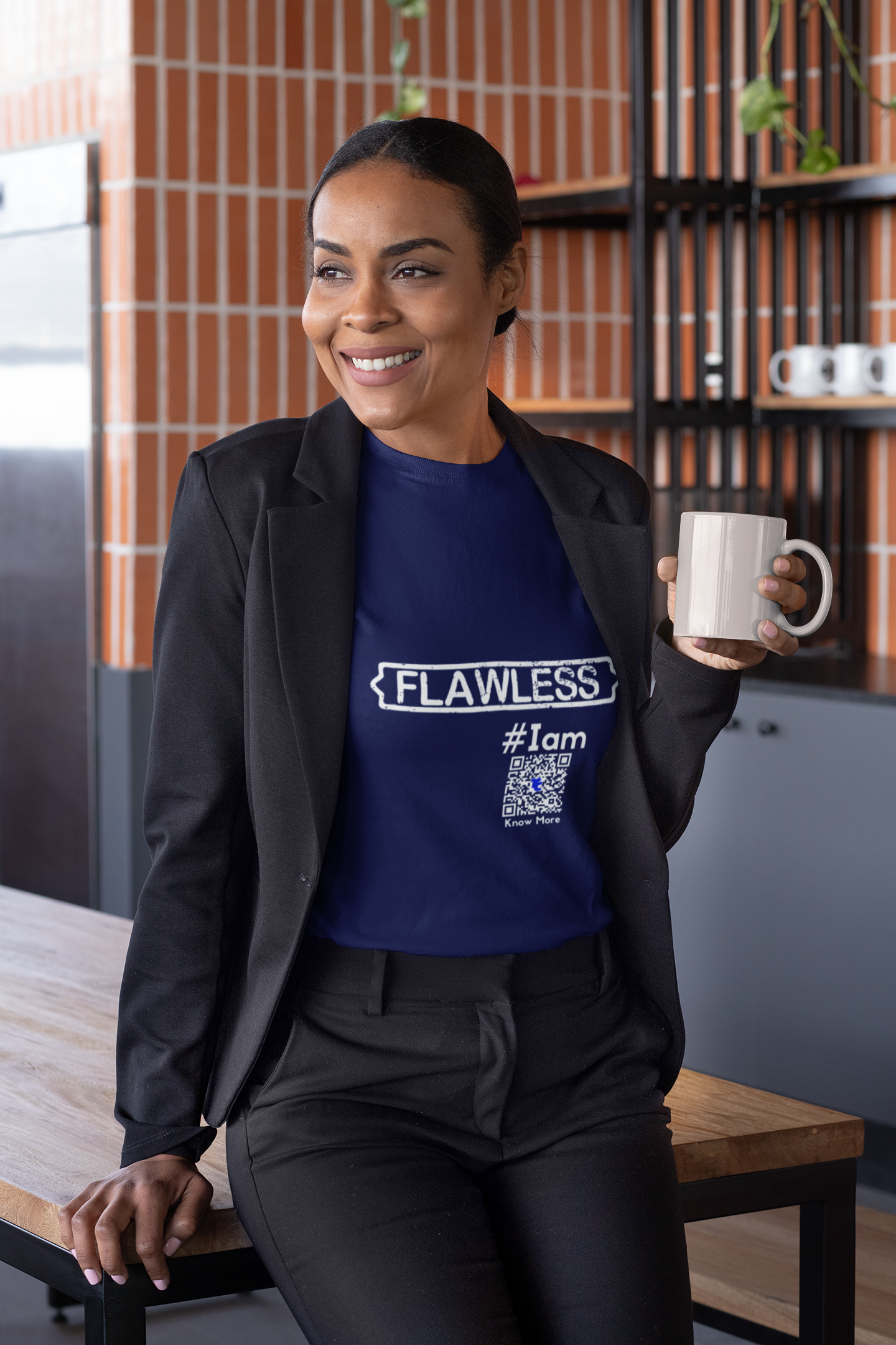 A smiling, dark skin, female presenting woman is sitting on the edge of a table. She is wearing a casual but professional looking black suit with a navy blue CLAIM It Tee™. The CLAIM™ reads {FLAWLESS} #Iam.