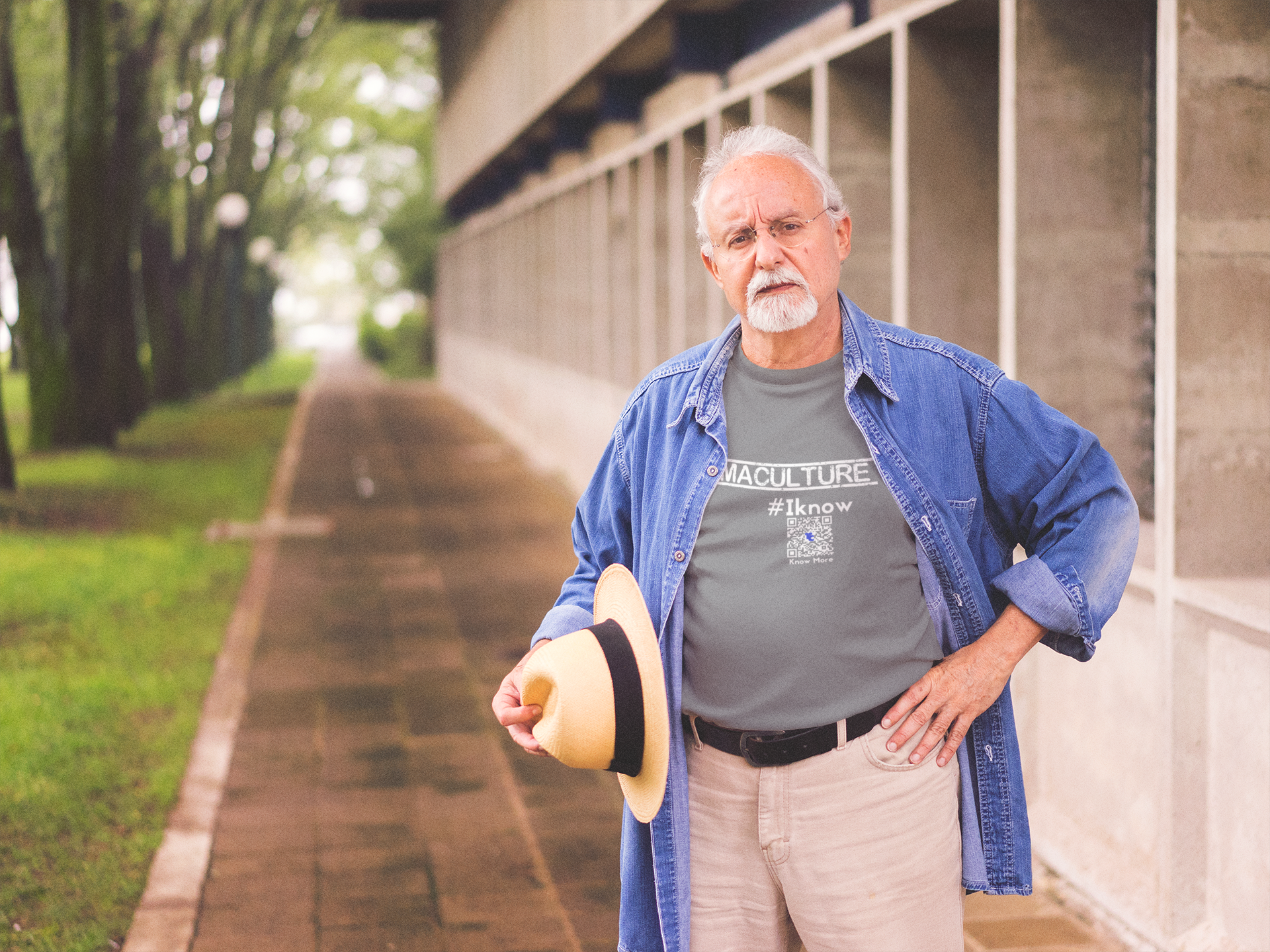 A male presenting person is standing outside with a straw hat in one hand & the other on their hip. The person, appears to be over 60 years in age & has gray hair with a gray goatee. They're wearing a gray CLAIM It Tee™ with a CLAIM™ that reads {PERMACULTURE} #Iknow.