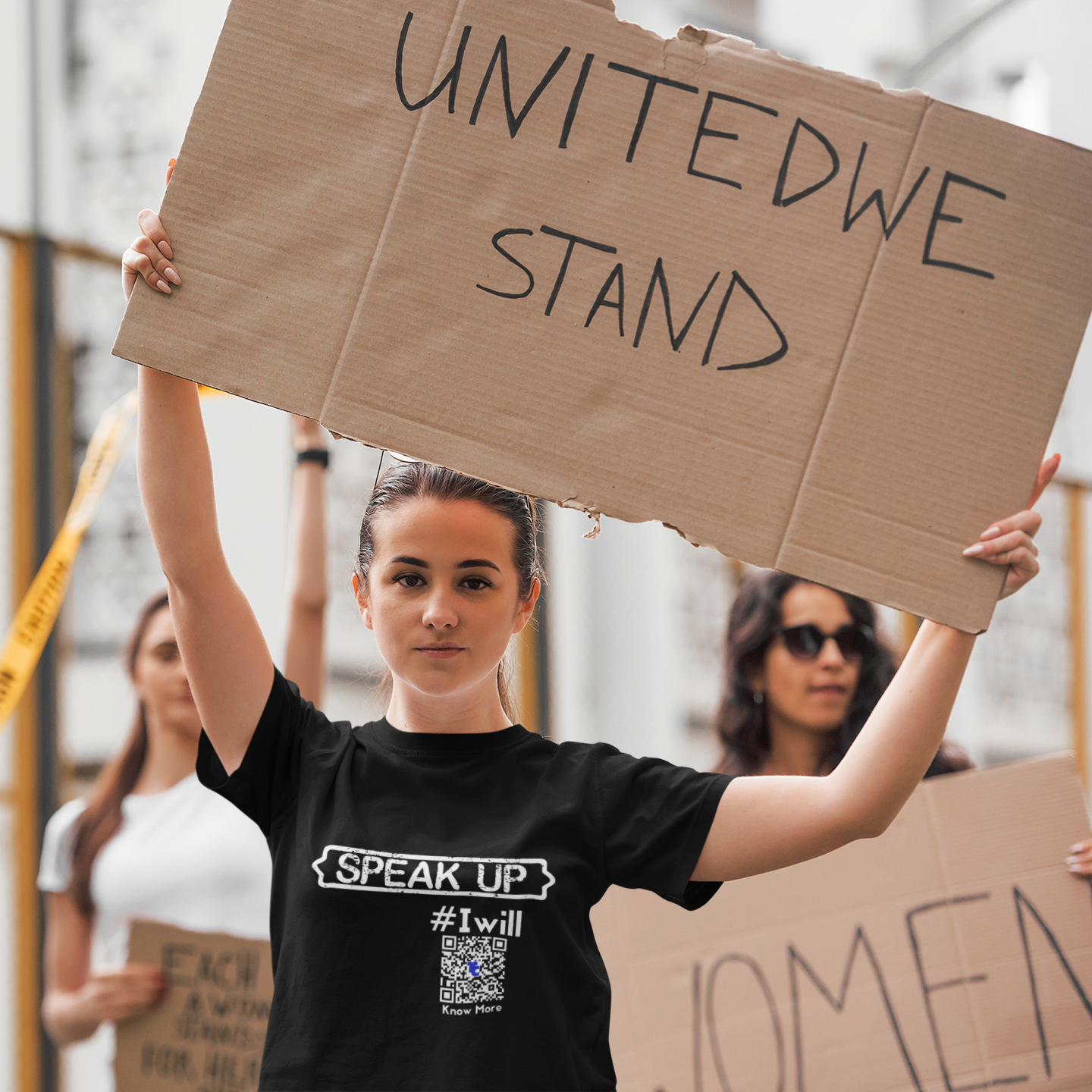 A female presenting person, that appears to be between the age of 16 and 20, is holding a large cardboard sign over their head that reads United We Stand. They appear to be marching with other female presenting people for women's rights. The person holding that sign is also wearing a black CLAIM It Tee™ with a CLAIM™ that reads {SPEAK UP} #Iwill. 