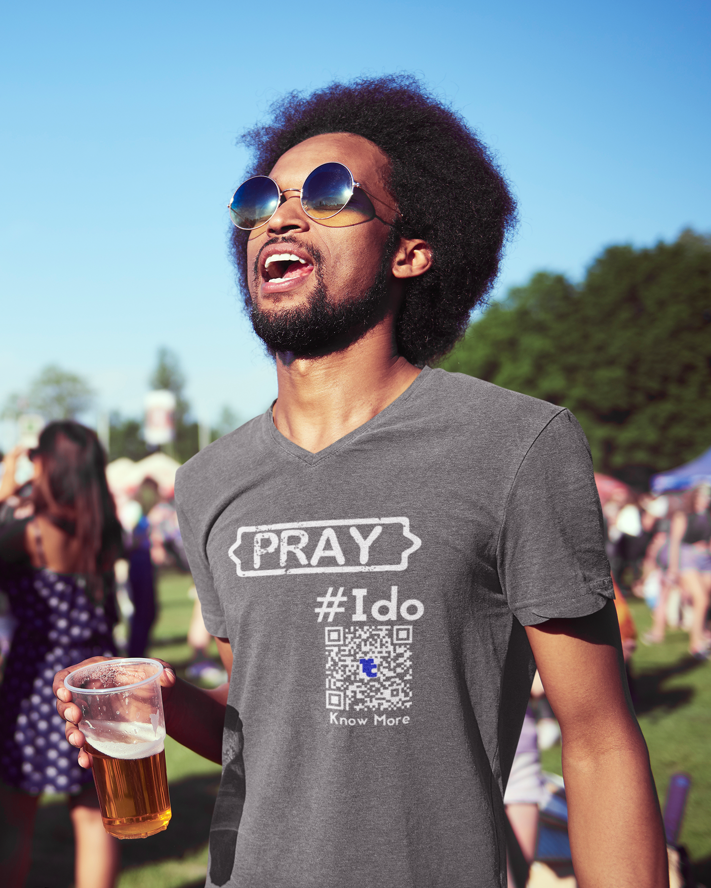 A male presenting person looks to be enjoying music & a beer while at an outdoor festival. The person is wearing a gray CLAIM It Tee™ with a CLAIM™ that reads {PRAY} #Ido. The QRcode, which is how people can access the share space™ for that shirt, is oversized & meant to be easy to scan. 