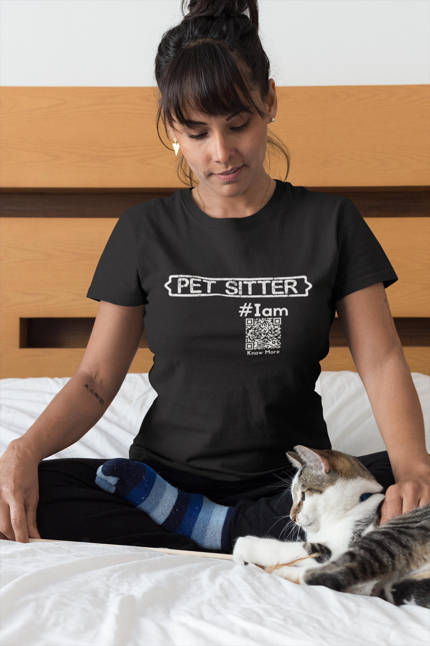 A female presenting person is sitting on a comfy looking bed with a cat. She is wearing a black CLAIM It Tee™ with a CLAIM™ that reads {PET SITTER} #Iam. The QRcode with the Share Space™ is visible. 